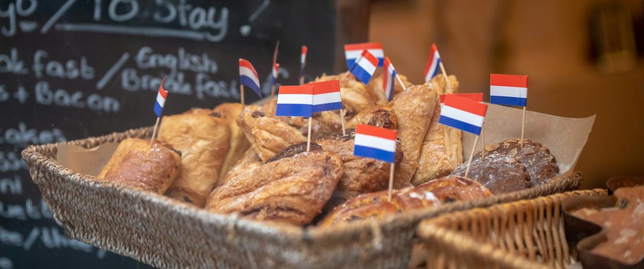 A wicker basket filled with assorted baked pastries, each decorated with a small Dutch flag toothpick, positioned in front of a chalkboard menu listing breakfast items.