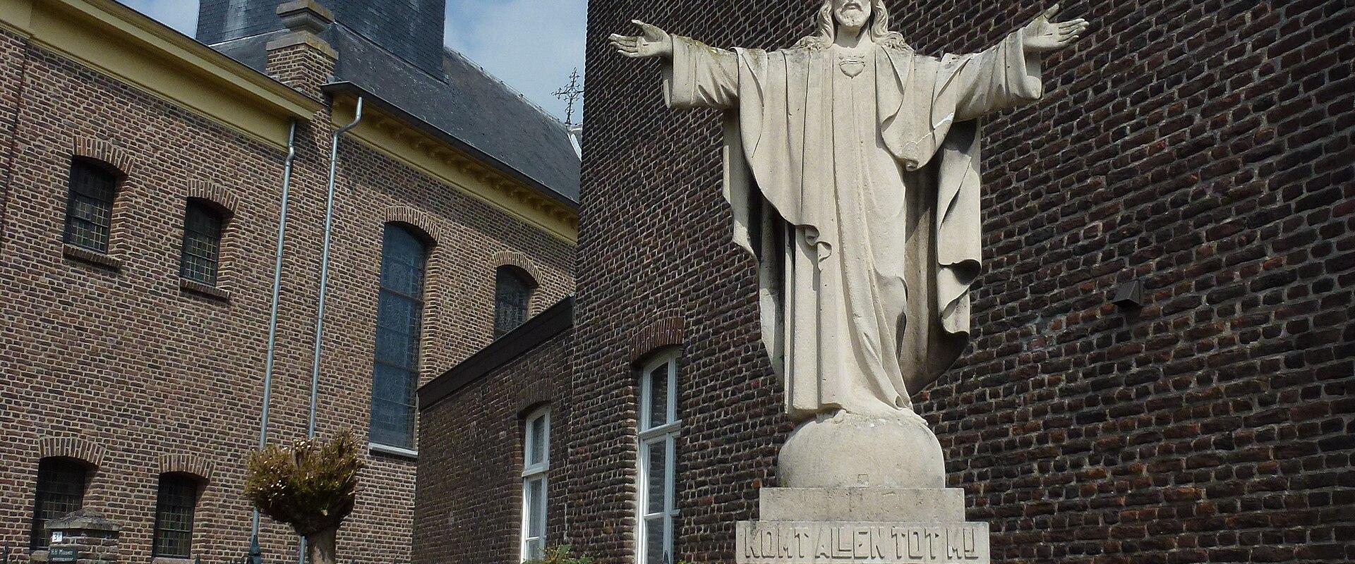 A stone statue of Jesus with outstretched arms stands on a pedestal inscribed with "KOMT ALLEN TOT MIJ" in front of a brick church building with a dark spire.