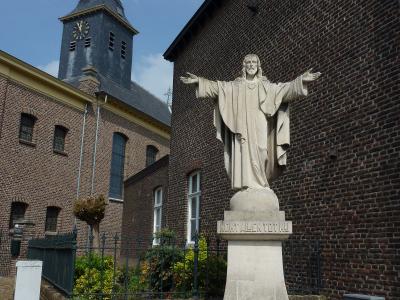 A stone statue of Jesus with outstretched arms stands on a pedestal inscribed with "KOMT ALLEN TOT MIJ" in front of a brick church building with a dark spire.