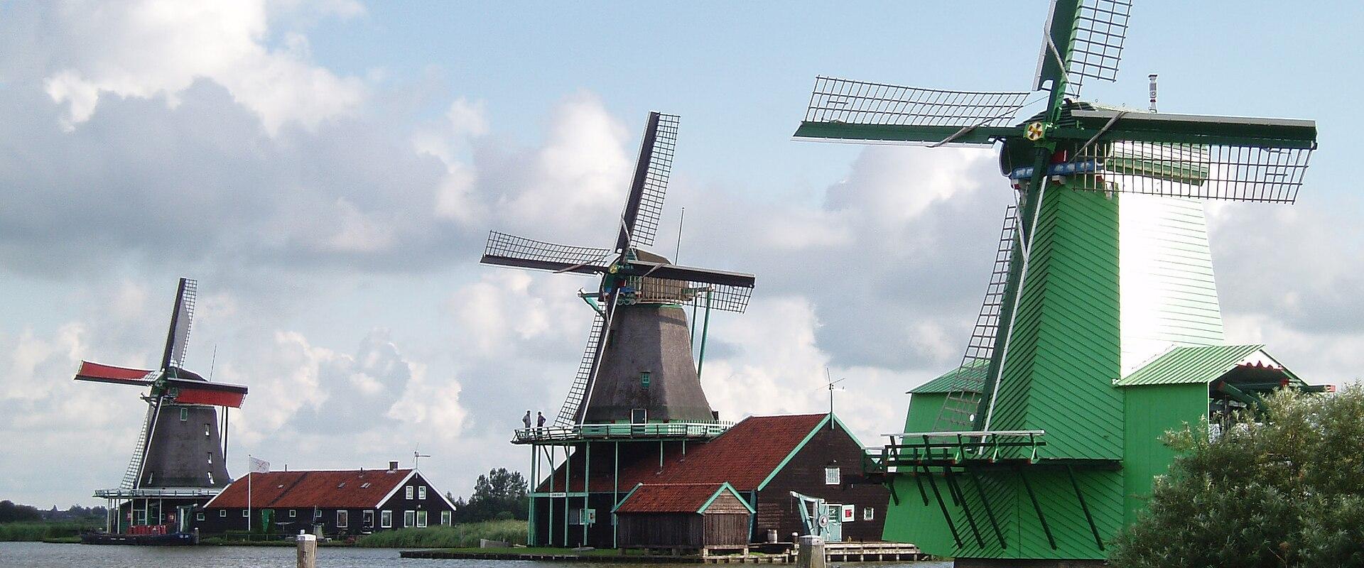 A view of three traditional Dutch windmills situated along a canal or riverbank, featuring a large green windmill in the foreground, a dark wooden windmill in the center, and a smaller windmill with red sails to the left.