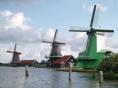 A view of three traditional Dutch windmills situated along a canal or riverbank, featuring a large green windmill in the foreground, a dark wooden windmill in the center, and a smaller windmill with red sails to the left.