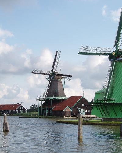 A view of three traditional Dutch windmills situated along a canal or riverbank, featuring a large green windmill in the foreground, a dark wooden windmill in the center, and a smaller windmill with red sails to the left.
