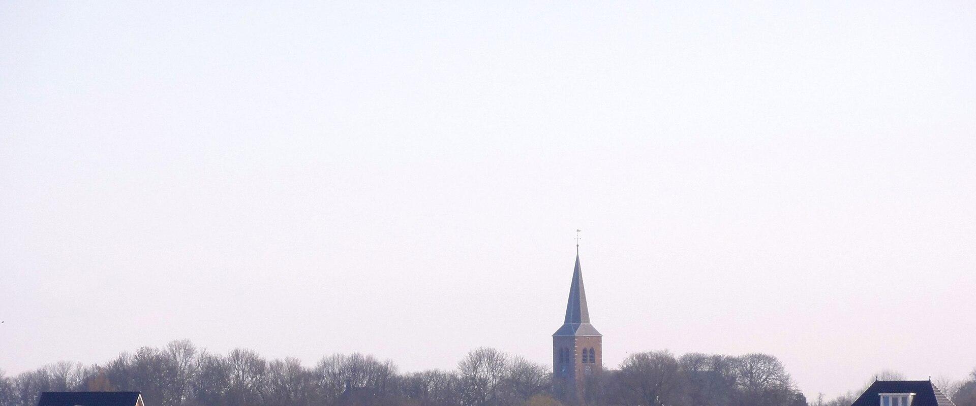 A landscape view of a Dutch village with a prominent church spire rising above a line of trees and houses, seen across a plowed field in the foreground.
