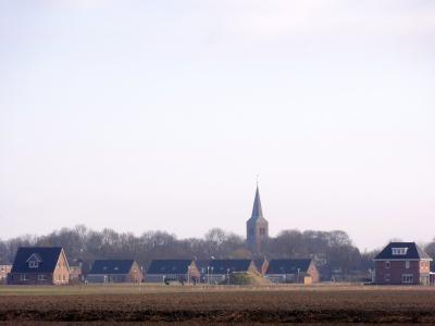 A landscape view of a Dutch village with a prominent church spire rising above a line of trees and houses, seen across a plowed field in the foreground.