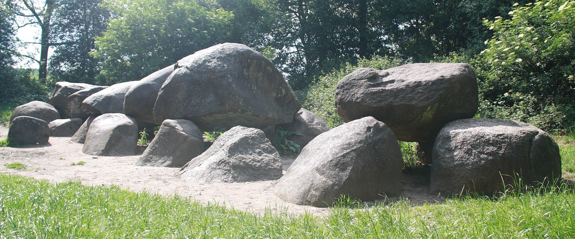 A low-angle photograph of a large, prehistoric megalithic tomb (dolmen) constructed from massive grey boulders, situated on a sandy clearing surrounded by green grass and dense trees.