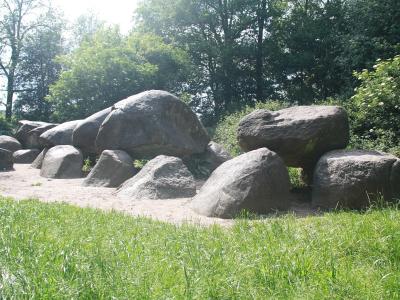 A low-angle photograph of a large, prehistoric megalithic tomb (dolmen) constructed from massive grey boulders, situated on a sandy clearing surrounded by green grass and dense trees.