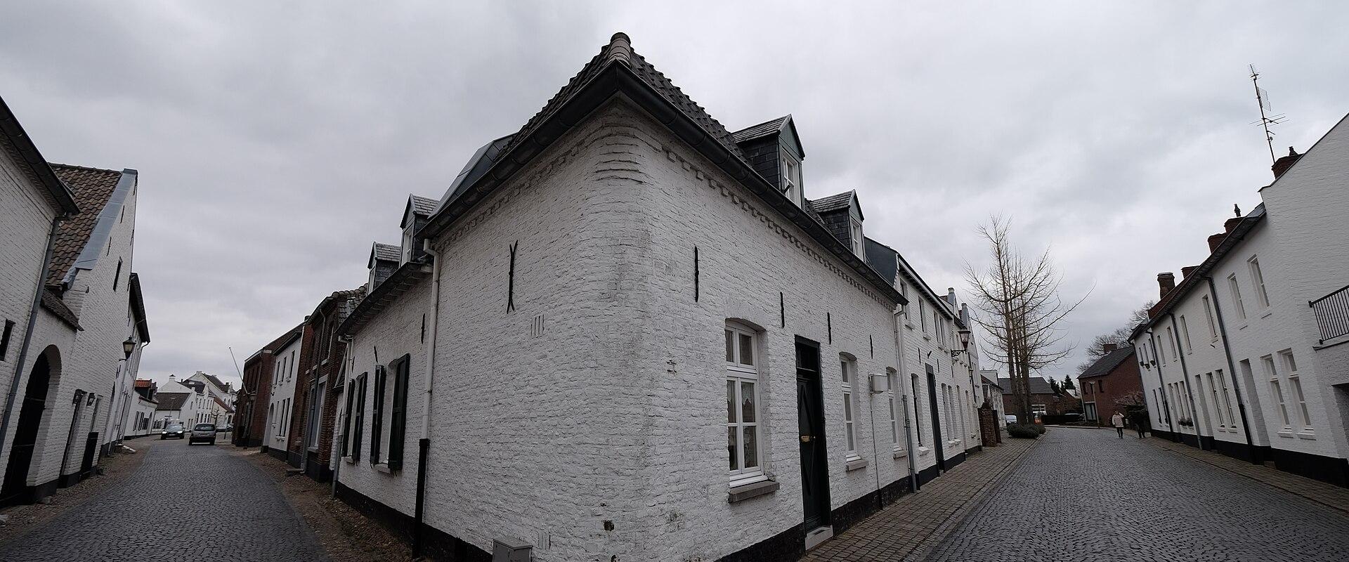 A street scene showing a prominent white-washed brick house at a fork in a cobblestone road, flanked by other traditional buildings under an overcast sky.