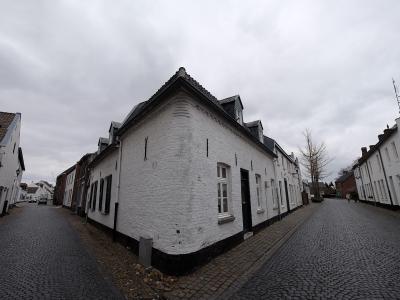 A street scene showing a prominent white-washed brick house at a fork in a cobblestone road, flanked by other traditional buildings under an overcast sky.
