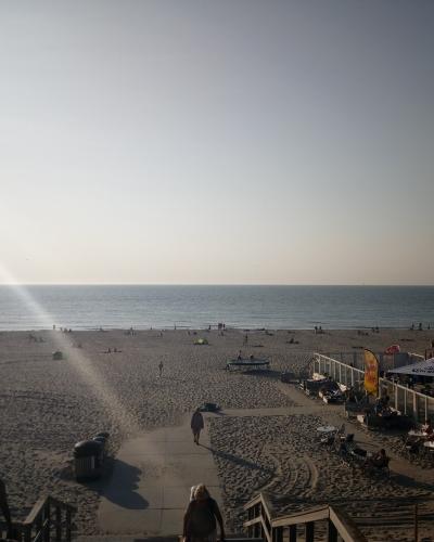 A wide view of a sandy beach with the North Sea in the background, featuring a wooden boardwalk, beachgoers, and a white building with outdoor seating on the right side.