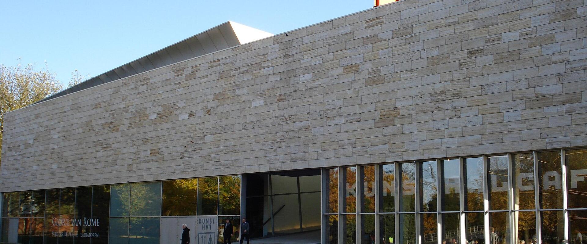 The image shows the exterior of a modern building with a stone facade and large glass windows, featuring signage for an exhibition titled "Oude Van Rome" and the word "KUNSTHAL" visible on the glass.