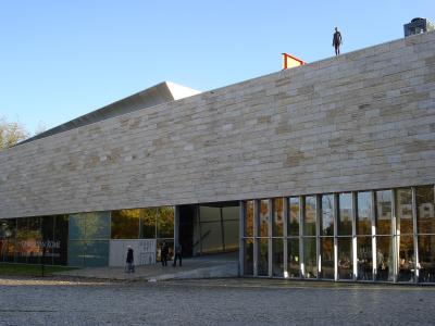 The image shows the exterior of a modern building with a stone facade and large glass windows, featuring signage for an exhibition titled "Oude Van Rome" and the word "KUNSTHAL" visible on the glass.