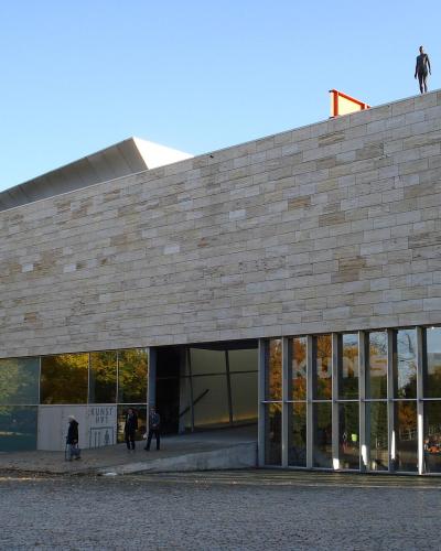 The image shows the exterior of a modern building with a stone facade and large glass windows, featuring signage for an exhibition titled "Oude Van Rome" and the word "KUNSTHAL" visible on the glass.