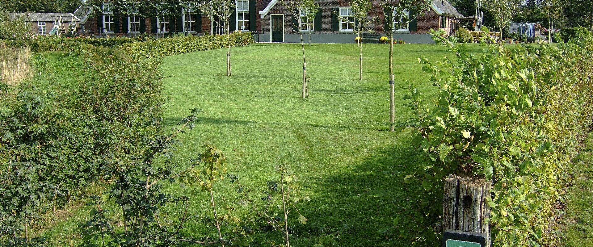 A view of a traditional Dutch brick farmhouse with a thatched roof, set behind a green lawn, with a green trail marker sign (labeled '5' and '5A') in the foreground.