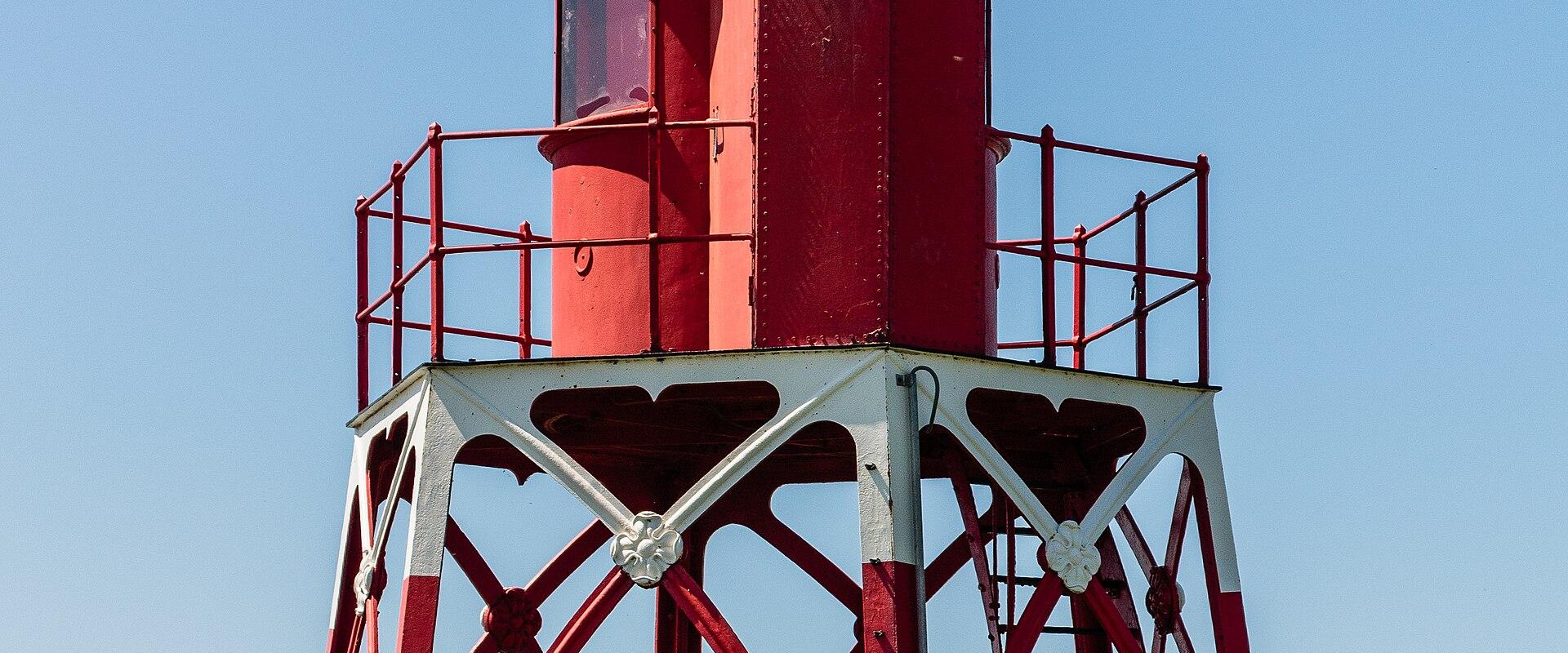 A red and white metal lighthouse structure standing on a grassy dike against a clear blue sky.