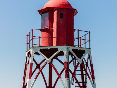 A red and white metal lighthouse structure standing on a grassy dike against a clear blue sky.