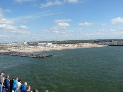 A group of people stand on a boat, looking out at a sandy beach lined with white tents and a large building, under a blue sky with scattered clouds.