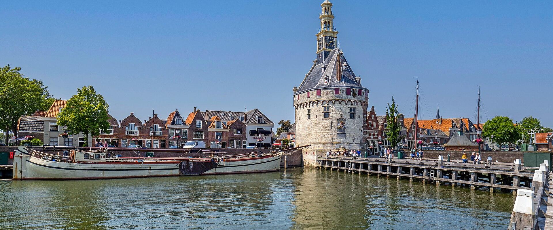 A view of the Hoofdtoren (Main Tower) in Hoorn, Netherlands, standing at the edge of a canal with a traditional boat moored nearby and historic buildings in the background.