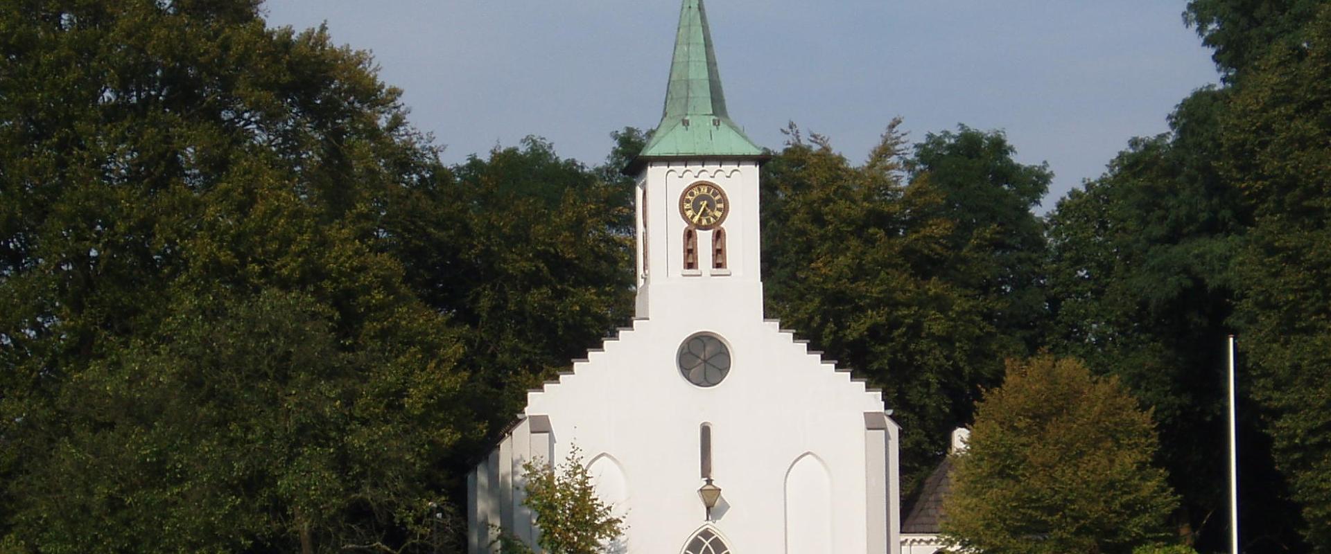 A white church with a green spire stands in a grassy field, surrounded by trees, with a clock face visible on its tower.