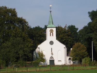 A white church with a green spire stands in a grassy field, surrounded by trees, with a clock face visible on its tower.