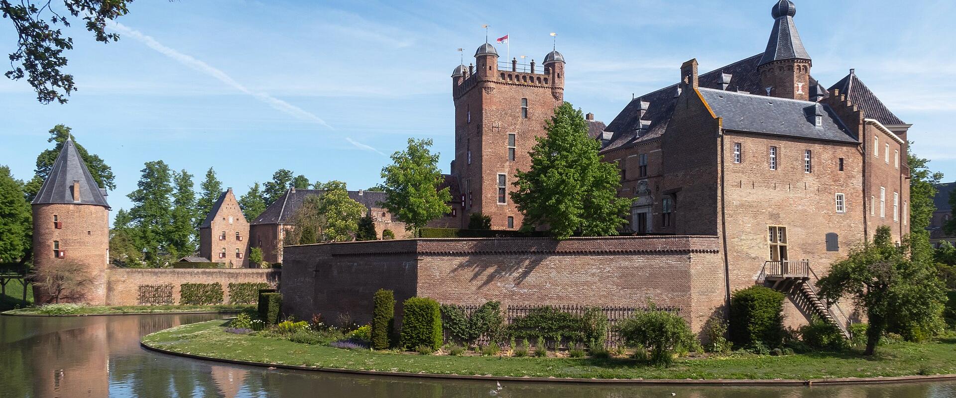 A historic brick castle with a moat, surrounded by greenery, under a clear blue sky.