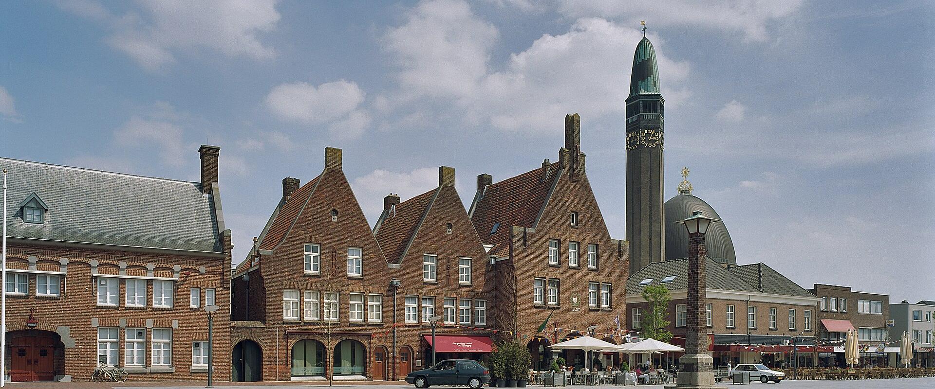 A brick town square featuring a row of historic buildings with stepped gables and a prominent church tower with a green spire and clock face in the background.