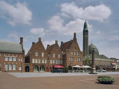 A brick town square featuring a row of historic buildings with stepped gables and a prominent church tower with a green spire and clock face in the background.