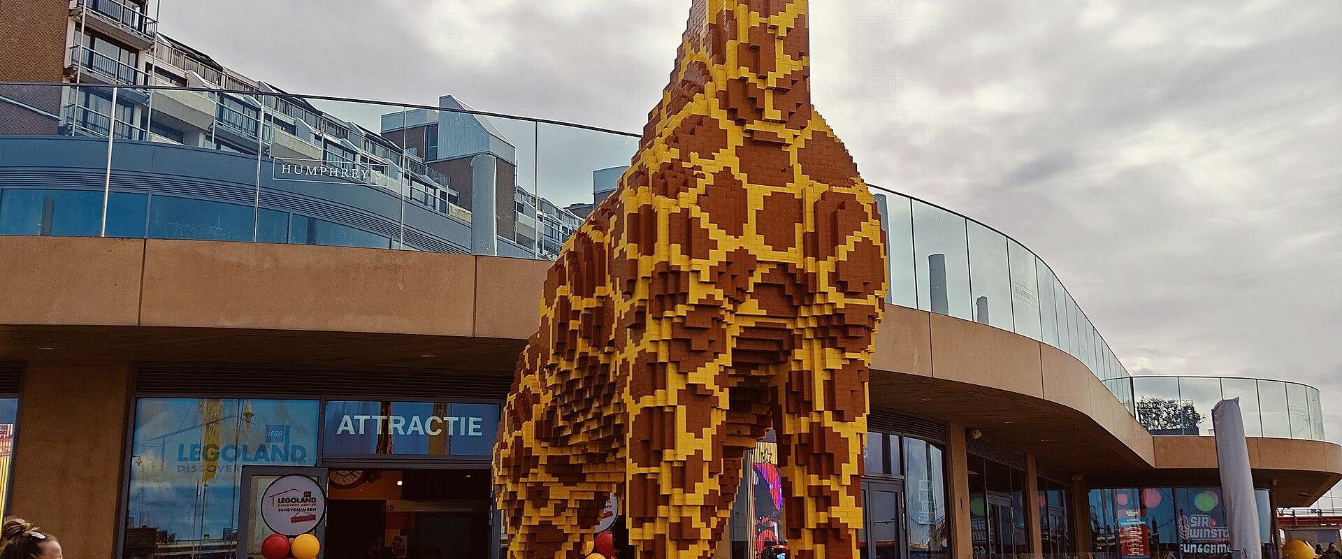 A large yellow and brown LEGO giraffe statue stands on a paved plaza in front of a modern building entrance with glass walls and signage reading "LEGOLAND" and "ATTRACTIE".