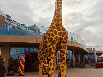 A large yellow and brown LEGO giraffe statue stands on a paved plaza in front of a modern building entrance with glass walls and signage reading "LEGOLAND" and "ATTRACTIE".