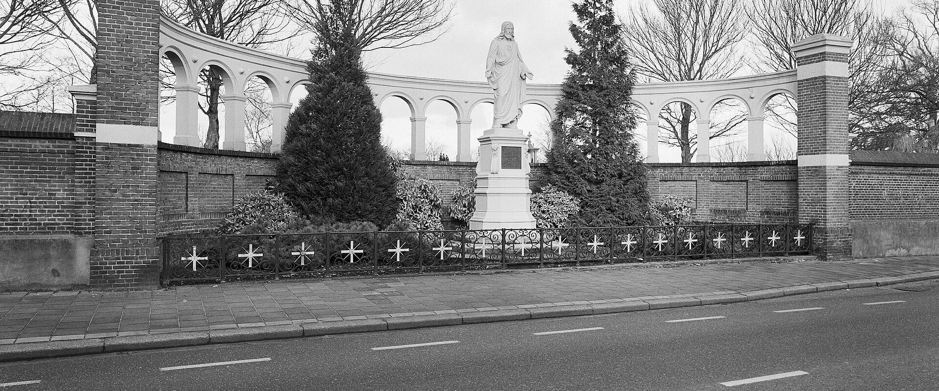 A black and white photograph showing a curved white colonnade wall with a statue of a robed figure on a pedestal in the center, flanked by brick pillars and a low iron fence with crosses, situated next to a paved road.