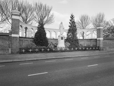 A black and white photograph showing a curved white colonnade wall with a statue of a robed figure on a pedestal in the center, flanked by brick pillars and a low iron fence with crosses, situated next to a paved road.