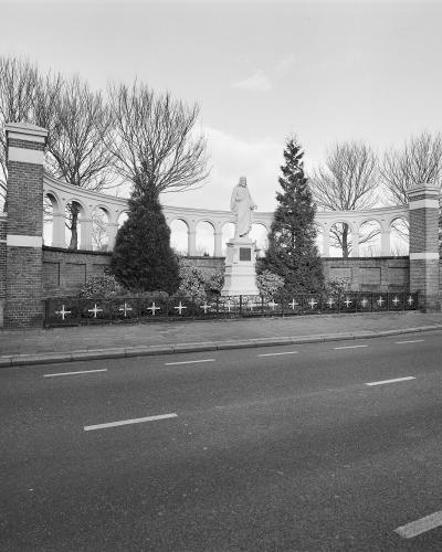 A black and white photograph showing a curved white colonnade wall with a statue of a robed figure on a pedestal in the center, flanked by brick pillars and a low iron fence with crosses, situated next to a paved road.
