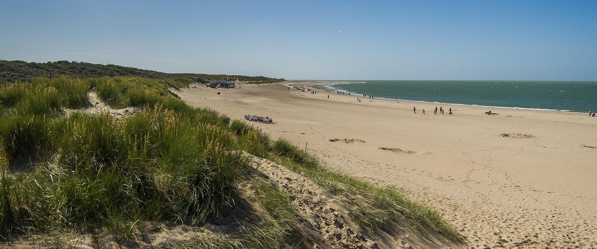 A wide, sandy beach scene with a blue sky, showing a few people on the sand, dune vegetation in the foreground, and a small structure (possibly a beach pavilion or lifeguard station) near the water's edge.