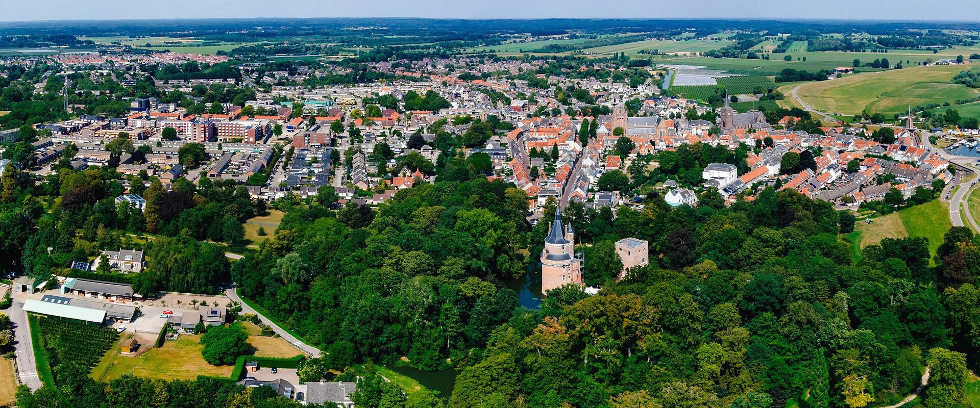 An aerial panoramic view of a Dutch town featuring a prominent castle with a conical-roofed tower and a square keep situated in a wooded park area, with the town's red-roofed buildings and surrounding flat countryside visible in the background.