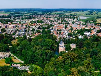 An aerial panoramic view of a Dutch town featuring a prominent castle with a conical-roofed tower and a square keep situated in a wooded park area, with the town's red-roofed buildings and surrounding flat countryside visible in the background.