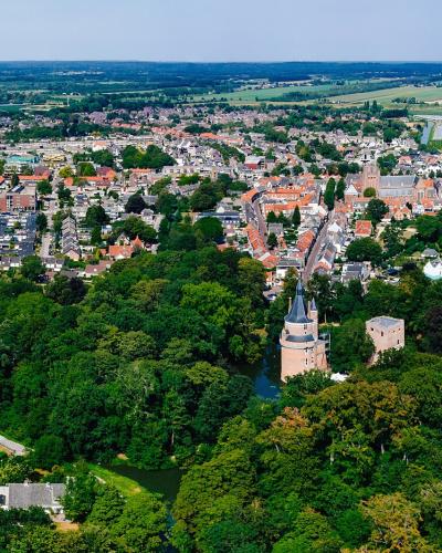An aerial panoramic view of a Dutch town featuring a prominent castle with a conical-roofed tower and a square keep situated in a wooded park area, with the town's red-roofed buildings and surrounding flat countryside visible in the background.