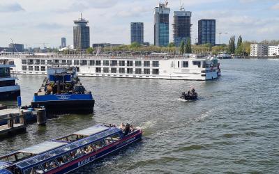 A Blue Boat tour vessel navigates a waterway with a large white river cruise ship docked nearby, set against a backdrop of modern high-rise buildings under construction and a control tower.