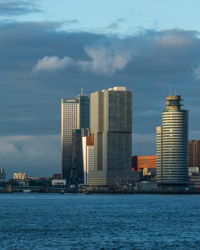 A view of the Maas river in Rotterdam featuring a Spido tour boat, the Erasmus Bridge in the distance, and the city skyline including the De Rotterdam skyscraper.