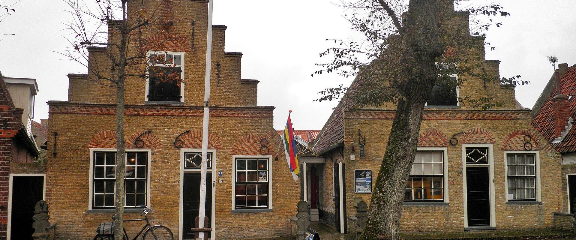 A traditional Dutch brick building with stepped gables, a flagpole, a bicycle, and a sandwich board sign on a cobblestone street.
