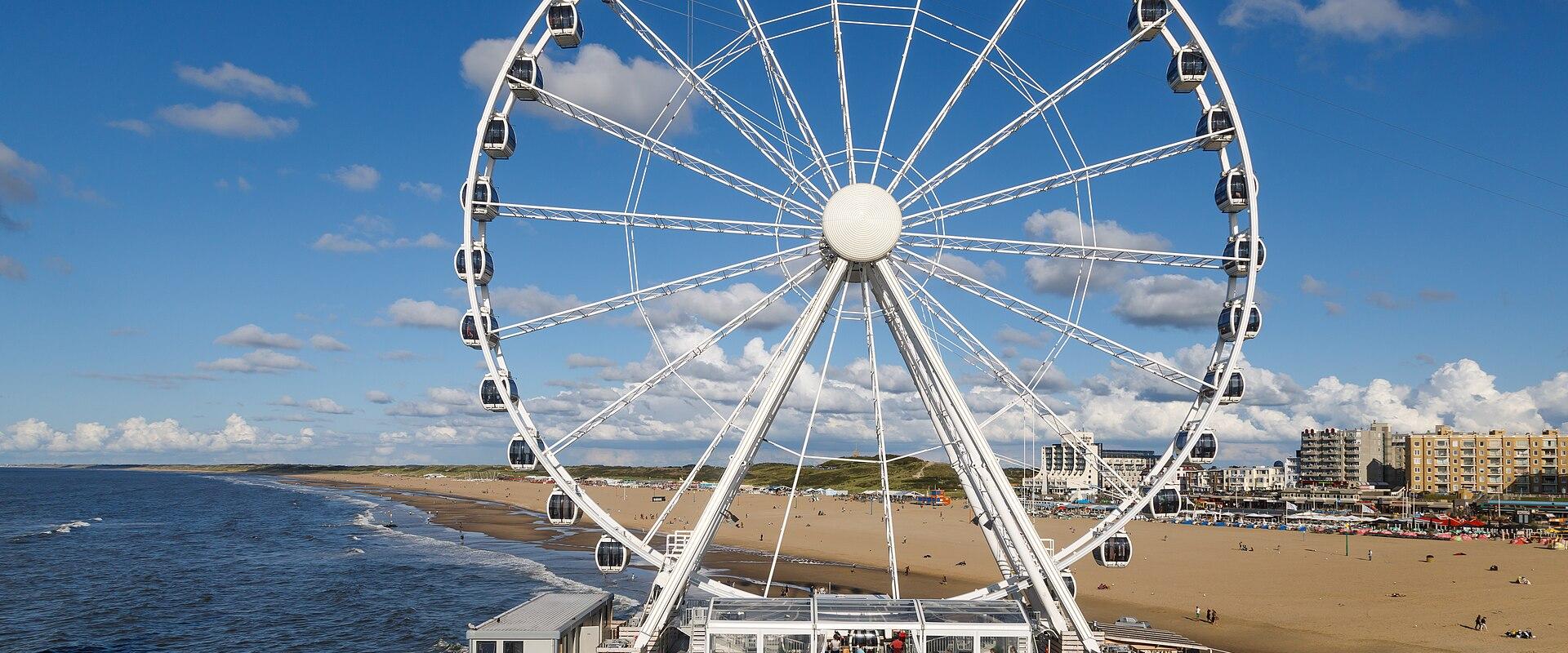 A large white Ferris wheel situated on a pier extending into the sea, with a sandy beach and coastal buildings visible in the background under a blue sky.