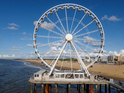 A large white Ferris wheel situated on a pier extending into the sea, with a sandy beach and coastal buildings visible in the background under a blue sky.