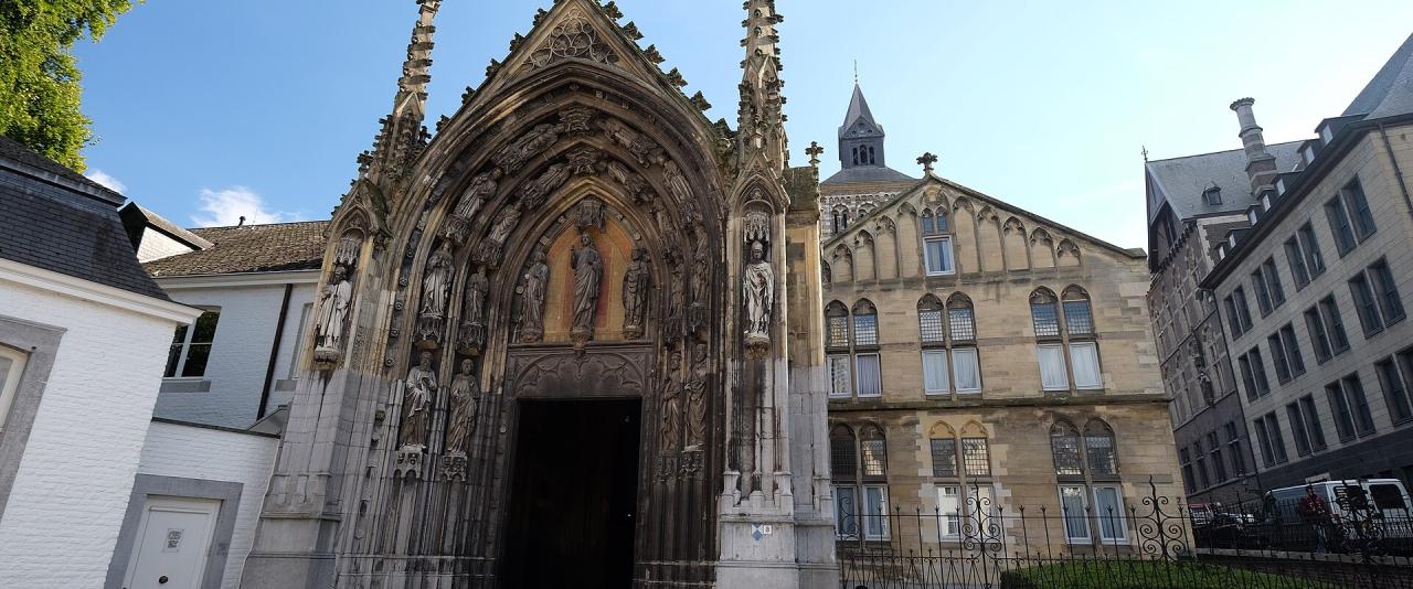 1. The image shows the ornate Gothic north portal of the Basilica of St. Servatius in Maastricht, Netherlands, with its detailed stone carvings, statues, and spires, set against a clear blue sky with adjacent historic buildings visible.