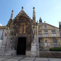1. The image shows the ornate Gothic north portal of the Basilica of St. Servatius in Maastricht, Netherlands, with its detailed stone carvings, statues, and spires, set against a clear blue sky with adjacent historic buildings visible.