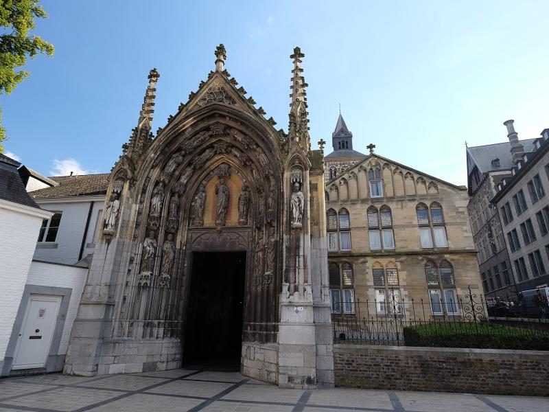 1. The image shows the ornate Gothic north portal of the Basilica of St. Servatius in Maastricht, Netherlands, with its detailed stone carvings, statues, and spires, set against a clear blue sky with adjacent historic buildings visible.