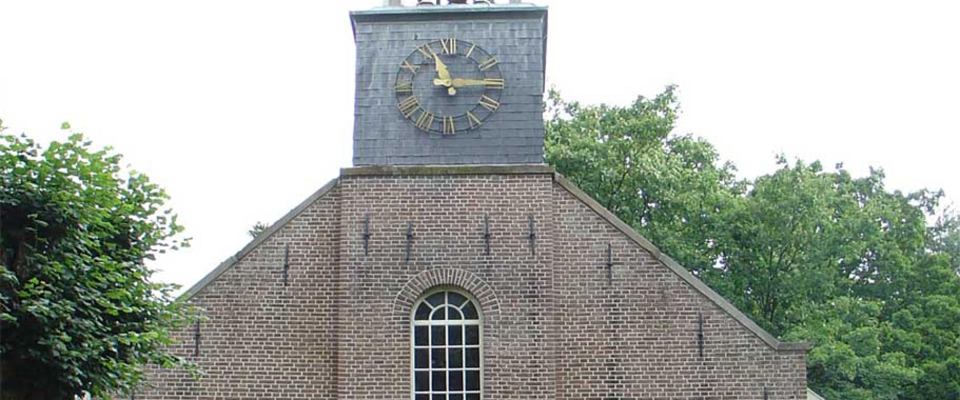 A front-facing photograph of a brick church with a central clock tower and spire, featuring arched windows and a plaque above the entrance.