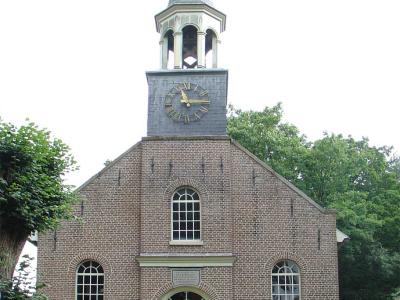 A front-facing photograph of a brick church with a central clock tower and spire, featuring arched windows and a plaque above the entrance.