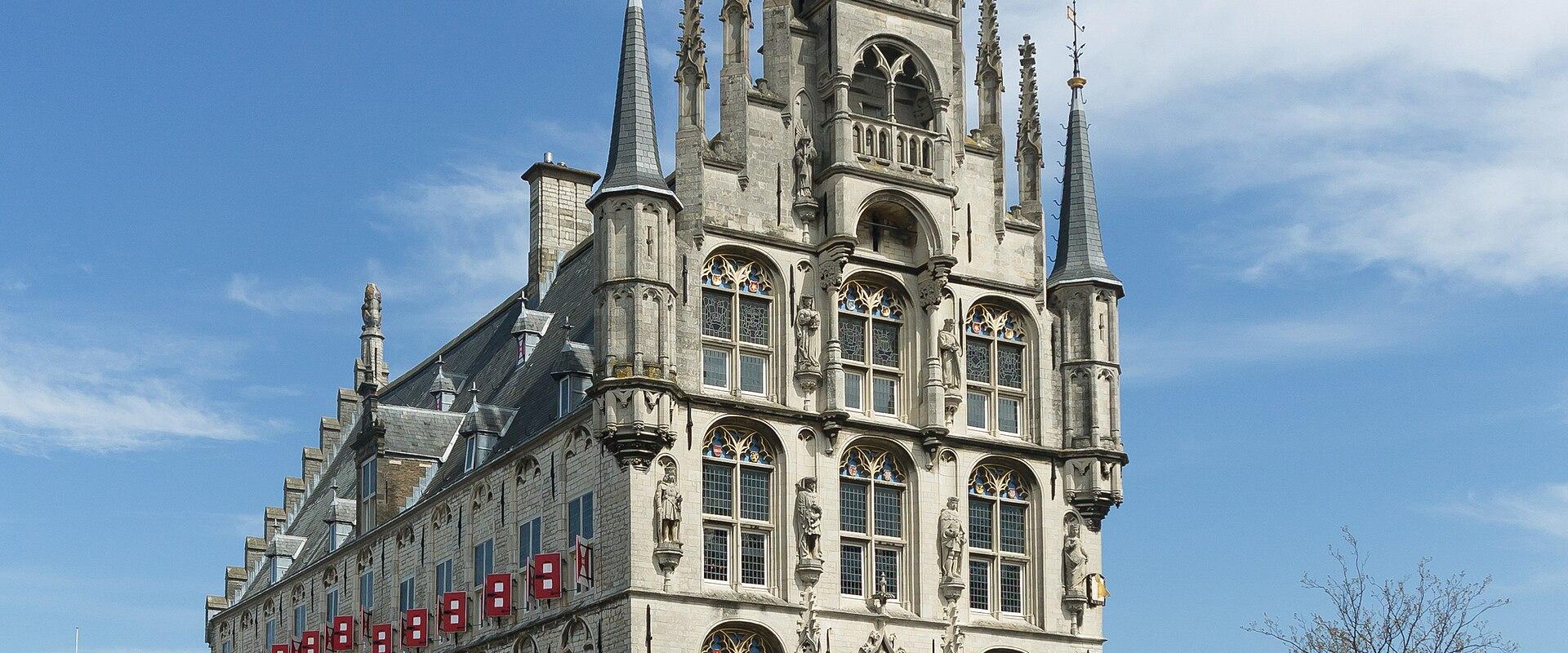 A large, ornate Gothic-style city hall with a tall central spire and stepped gable, situated on a paved public square under a blue sky with scattered clouds.