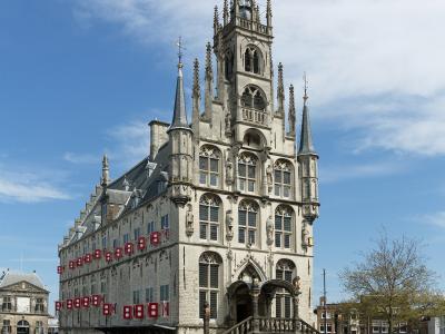 A large, ornate Gothic-style city hall with a tall central spire and stepped gable, situated on a paved public square under a blue sky with scattered clouds.