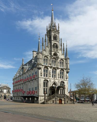 A large, ornate Gothic-style city hall with a tall central spire and stepped gable, situated on a paved public square under a blue sky with scattered clouds.