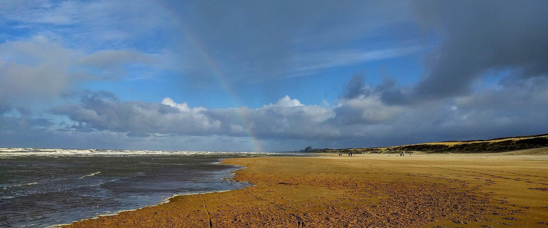 A wide sandy beach along the North Sea coast with a rainbow arching across a cloudy blue sky, dunes on the right, and a few distant figures walking along the shore.