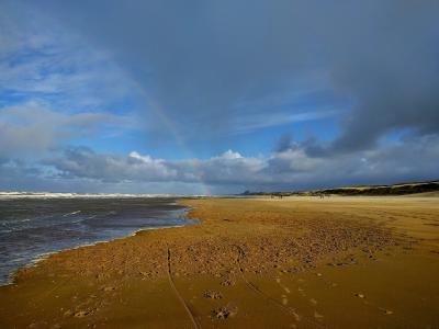 A wide sandy beach along the North Sea coast with a rainbow arching across a cloudy blue sky, dunes on the right, and a few distant figures walking along the shore.